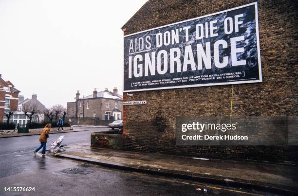 Woman pushes a stroller across an intersection , past a AIDS awareness campaign billboard on the wall of a house, South West London, England,...