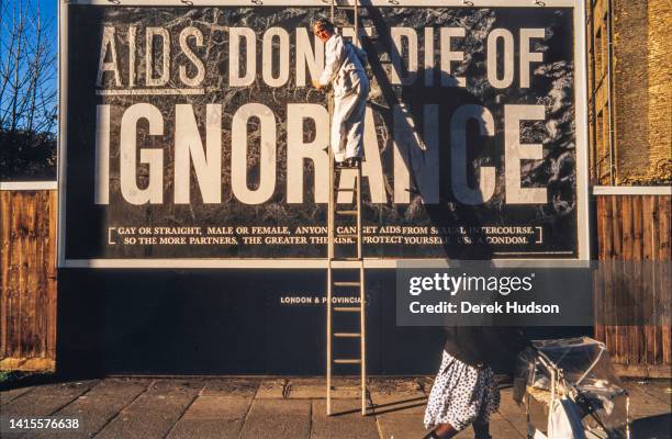 As a woman pushes a stroller past, an unidentified worker on a ladder pastes up an AIDS awareness campaign billboard, South West London, England,...