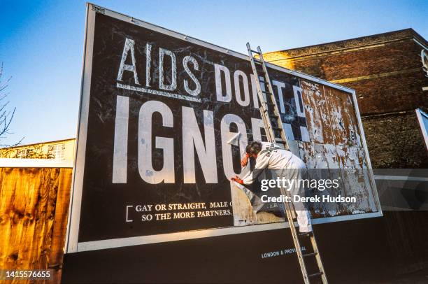 View of an unidentified worker on a ladder as he pastes up an AIDS awareness campaign billboard, South West London, England, December 1986. The...