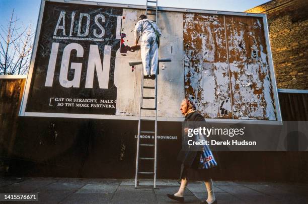 As a pedestrian passes by, an unidentified worker pastes up an AIDS awareness campaign billboard, South West London, England, December 1986. The...