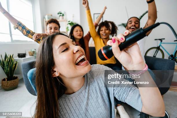 excited group of diverse millennial friends having fun playing karaoke together at home. young woman singing on video game. concept about friendship, home entertainment and people - jugendkultur sitzen konsole stock-fotos und bilder