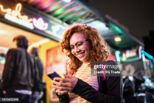 young woman using the mobile phone outdoors at night - food truck festival stock pictures, royalty-free photos & images