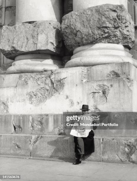 View of a man who is reading the newspaper 'Corriere della Sera', sat down on a step of the monumental Central Railway Station. Milan, the '50s.