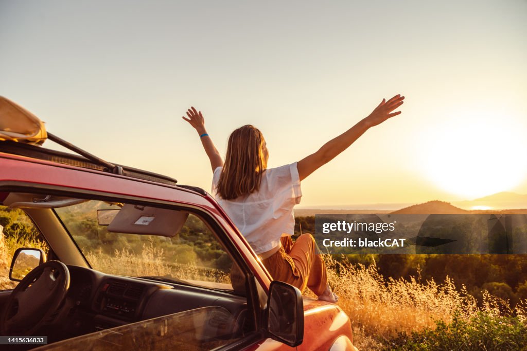Une jeune femme lève les bras assise sur la voiture et profite du coucher de soleil