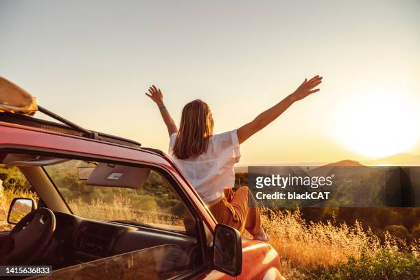 young woman arms raised sitting on the car and enjoying the sunset - road trip 個照片及圖片檔