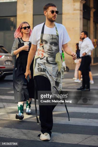 Guest wears black sunglasses, an apron with a face photograph print, white t-shirt, black pants, and cream Yeezy Foam Runner sneakers outside the...