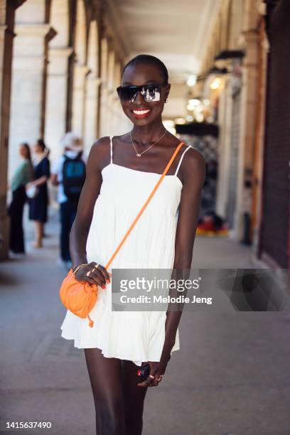 Model Nyagua Ruea wears black and purple square sunglasses, a white summer mini sleeveless dress, gold rings and accessories, and a small orange...