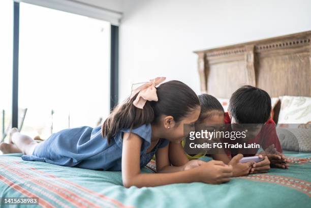 siblings using the mobile phone in the bed at home - modern family media call stockfoto's en -beelden