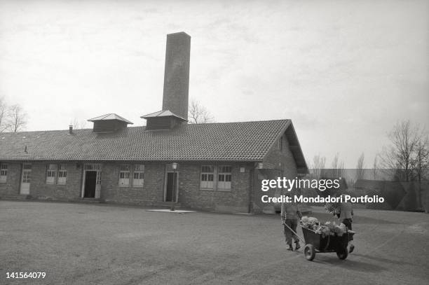 Man and a woman carry flowers in a wheelbarrow towards the former Dachau crematorium.