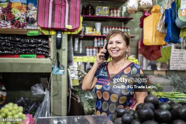 mature woman retail shop owner at her market store talking in a mobile phone - grocer stock pictures, royalty-free photos & images