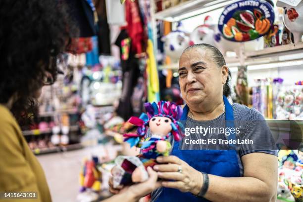 mujer medianamente adulta tomando muñeca mexicana de una mujer mayor en una tienda de regalos - artesanias mexicanas fotografías e imágenes de stock