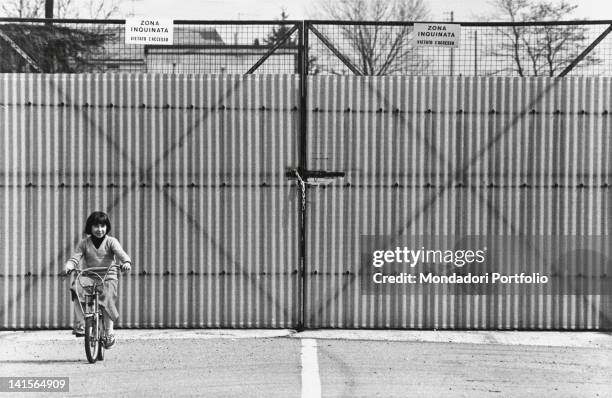 Young Italian girl riding her bike in front of the gate that delimits the area in Seveso that has been polluted the most with dioxin. Seveso, 1970s