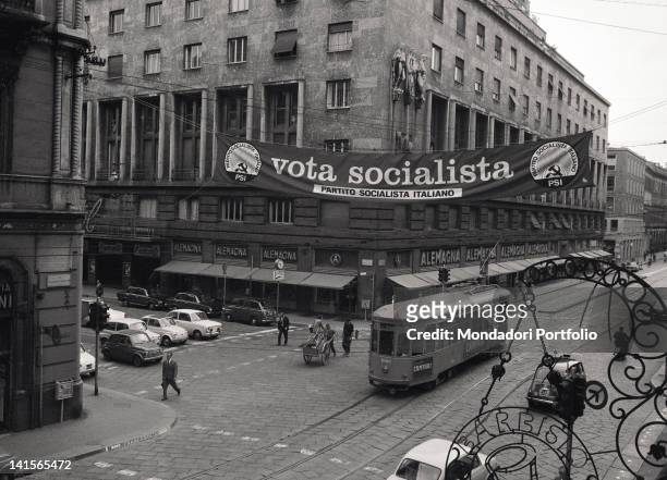 View of the crossing between Via Manzoni and Via Croce Rossa crossed by cars and trams, and surmounted by an election banner. Milan, 1970