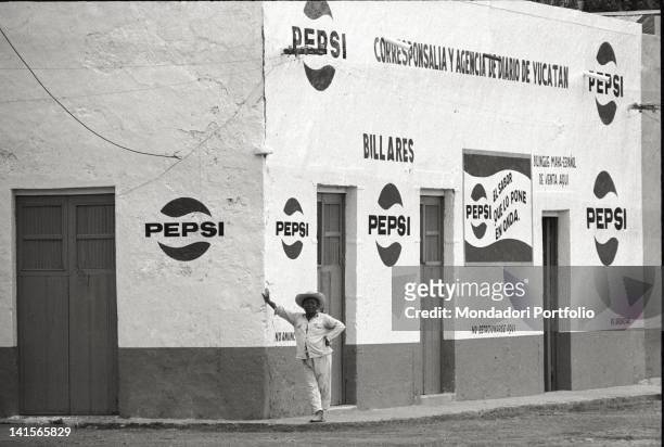 Mexican man, with a sombrero on his head, is leaning against a wall of a building which is almost full decorated with the logo of the American...