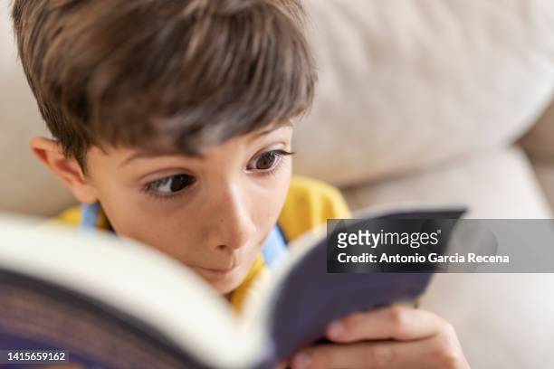 6 year old little boy reads a book on the sofa at home - boy funny face ストックフォトと画像