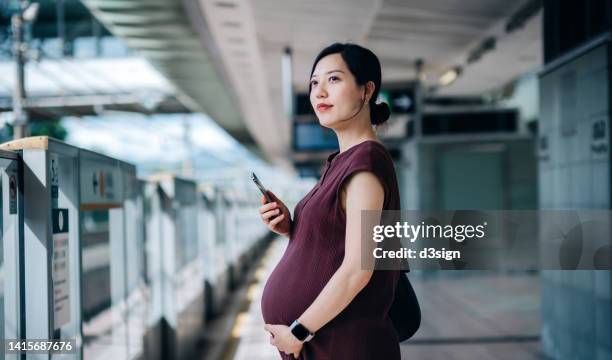 young asian pregnant woman using smartphone while commuting with subway in the city, gently touching her baby bump, waiting for train at the subway platform - fund fair imagens e fotografias de stock