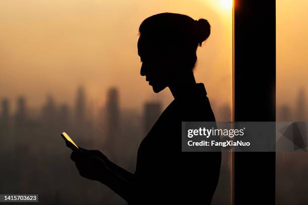 silhouette of young woman using smartphone next to window with cityscape - prostitutie stockfoto's en -beelden