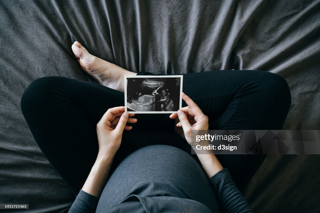 High angle shot of Asian pregnant woman holding an ultrasound scan photo in front of her baby bump, sitting on bed at home. Mother-to-be. Precious moment in life. Preparation for a new family member. Expecting a new life. Baby and new life concept
