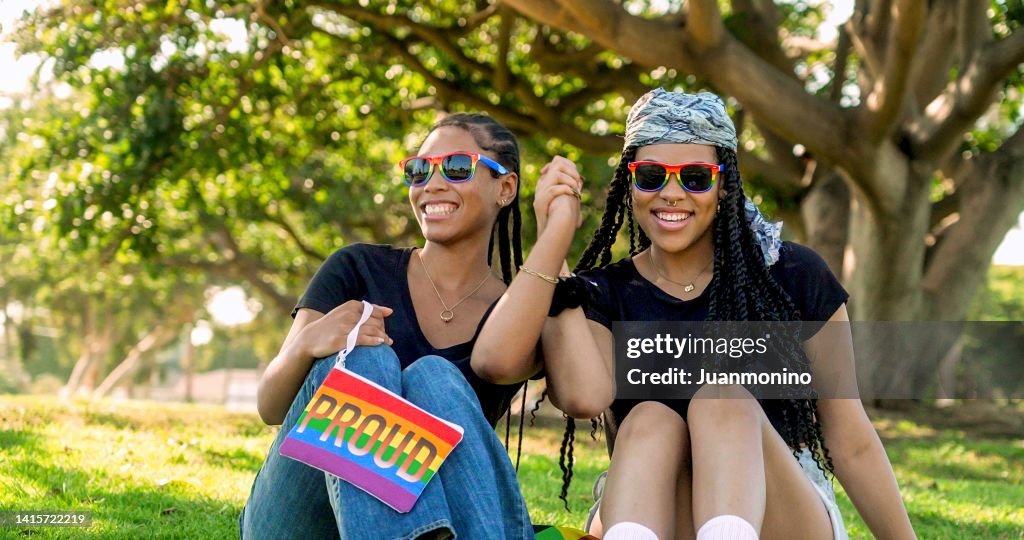 Afro-latinx young women sitting on the grass in a park holding hands looking at the camera