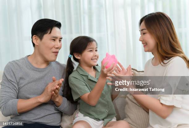 happy family saving money for future purchase, making reserve fund, planning budget. couple of parents and two little kids collecting cash, dropping coins into piggybank at home. financial education - asian family fall stock-fotos und bilder