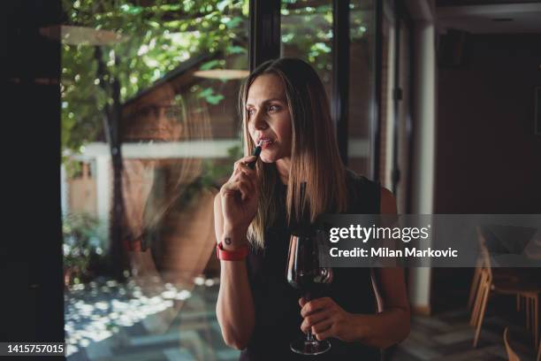 a beautiful woman is standing by the window holding a glass of wine and an electronic cigarette - hookah imagens e fotografias de stock