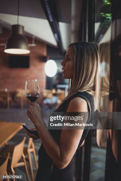 a beautiful woman is standing by the window holding a glass of wine and an electronic cigarette - hitech mod a 個照片及圖片檔