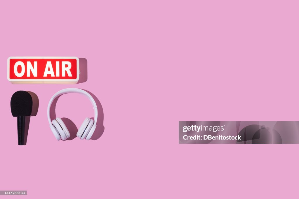 Lightbox turned on with the word 'On air', headphones and microphone with windscreen, on the left, on a pink background. Concept of radio, podcast, streaming, broadcast, journalism, reporting.