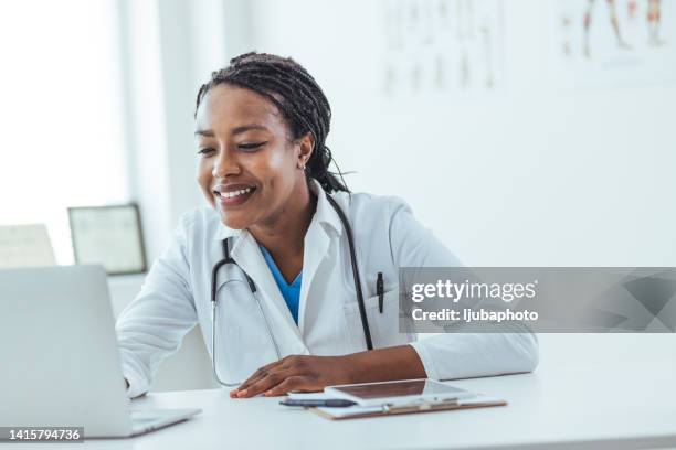 concentrated afro american woman doctor using laptop while sitting at workplace in medical clinic - stralende lach stockfoto's en -beelden