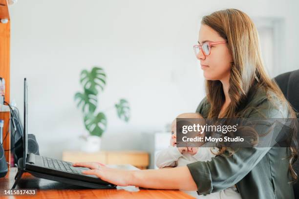 young mother with baby sitting at desk, working on laptop - working mother stock pictures, royalty-free photos & images