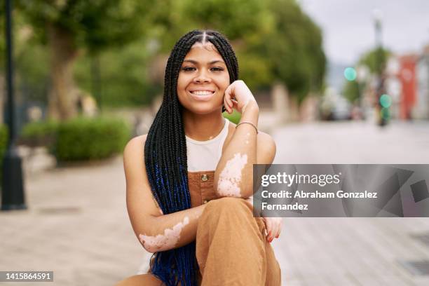 a happy young woman with vitiligo sitting on a sidewalk in the city - vitiligo stock-fotos und bilder