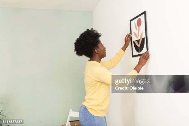 happy african american woman hanging a picture on the wall while moving in - hanging artwork stock pictures, royalty-free photos & images