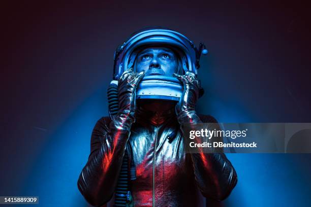 close up shot of astronaut cosmonaut dressed in gold jumpsuit and helmet, illuminated by blue and red lights, holding helmet and looking up. concept of exploration, space, planet, strange and alien. - alien arrival stock pictures, royalty-free photos & images