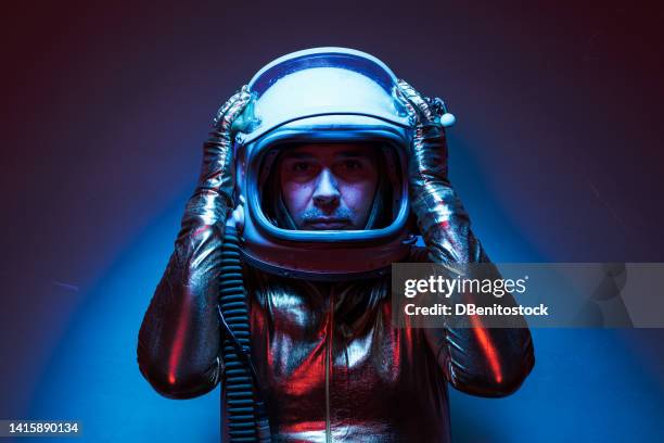 close shot of cosmonaut astronaut dressed in a gold jumpsuit and helmet, illuminated by blue and red lights, with his hands on his head. concept of exploration, space, planet, strange and alien. - astronaut helmet ストックフォトと画像