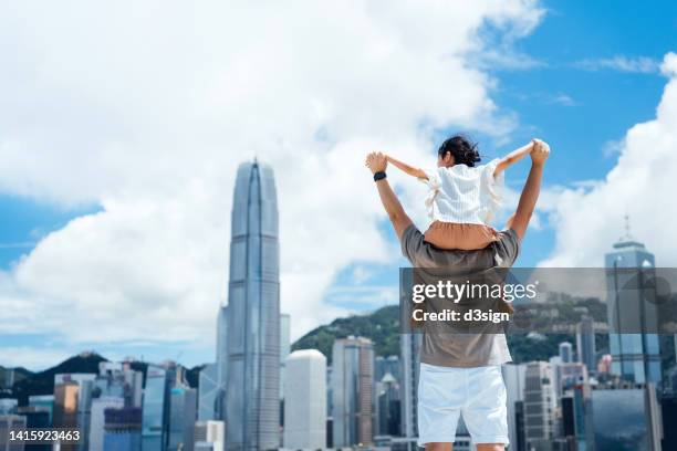 rear view of loving young asian father carrying little daughter on shoulders, looking over prosperous urban city skyline against the promenade of victoria harbour with blue sky. enjoying father and daughter bonding time. family love and lifestyle - piggy back stockfoto's en -beelden