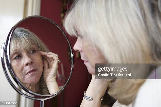 Woman aged 62 examines her face in the mirror three weeks after her facelift operation. The scars on her eyelids are fading but her face is still...