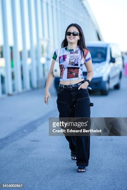 Guest wears black sunglasses, silver earrings, a silver chain pendant necklaces, a purple / white / blue short sleeves cropped t-shirt, a white matte...