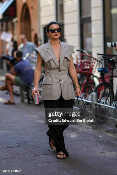 Guest wears black sunglasses, gold earrings, a pale gray shoulder-pads / buttoned / sleeveless blazer jacket, black large pants, black flip flop, a...