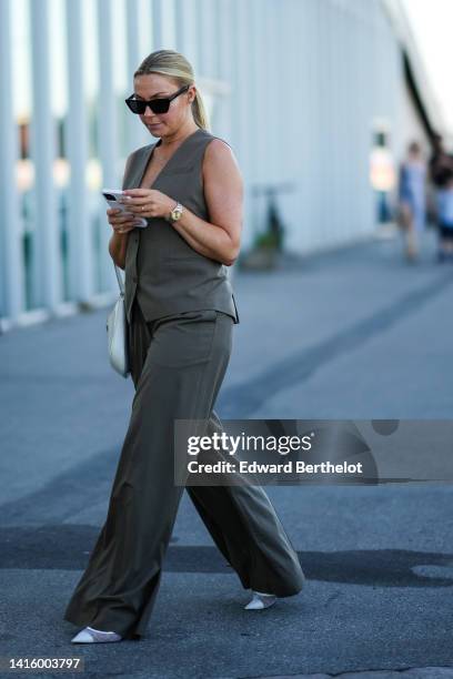 Guest wears black sunglasses, a gray sleeveless / buttoned gilet, dark gray large suit pants, a white shiny leather Cleo handbag from Prada, white...