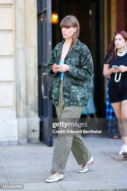 Guest wears silver earrings, a pale blue wrap / ruffled shoulder-off top, a dark green with black print pattern oversized bomber coat, a black shiny...