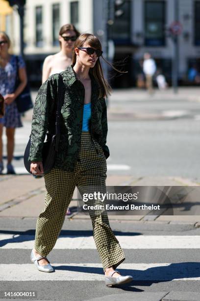 Guest wears silver earrings, a pale blue wrap / ruffled shoulder-off top, a dark green with black print pattern oversized bomber coat, a black shiny...