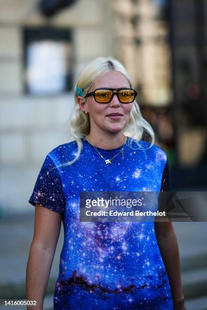 Guest wears black sunglasses, a blue studded hair clip, a blue and purple constellation print pattern t-shirt, a gold chain pendant necklace ,...