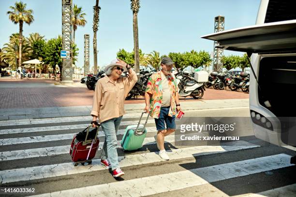 retired couple crossing street with wheeled luggage - coach bus 個照片及圖片檔