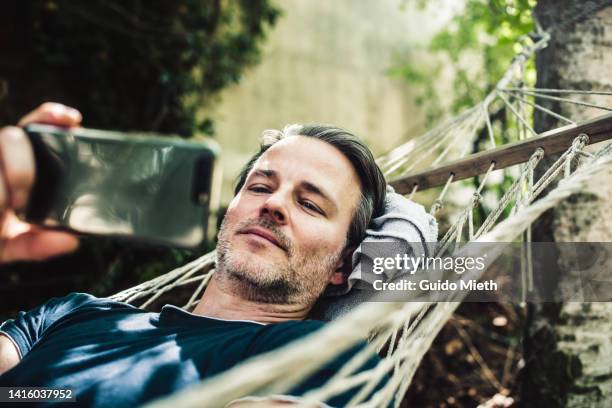 mature man lying in hammock with smart phone outdoor in sunlight. - german films stock pictures, royalty-free photos & images