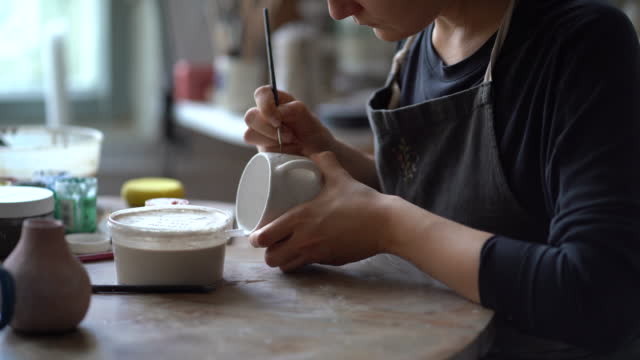 Craftswoman in black apron dips brush into can with paint to draw pattern on white handmade mug