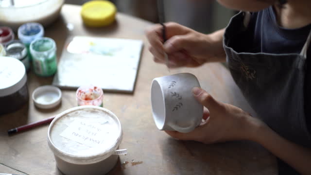 Female artisan in black apron paints patterns with brush on white ceramic mug sitting at table