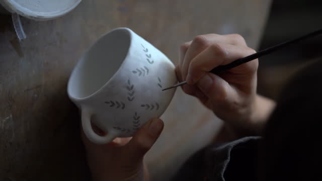 Woman enjoys handmade pottery craft painting creative pattern on white ceramic mug with brush