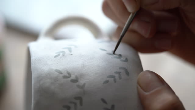 Woman draws pattern with dark paint on white ceramic mug using brush on blurred background