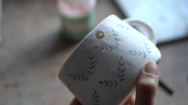 Woman draws pattern with dark paint on white ceramic mug using brush on blurred backgroundx