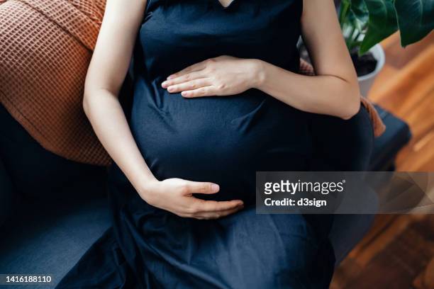 cropped shot of asian pregnant woman gently touching her baby bump, relaxing on the sofa at home. mother-to-be. expecting a new life. love and bonding concept - baby touching belly fotografías e imágenes de stock