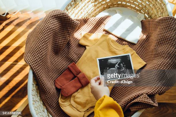 cropped hand of an expectant mother holding an ultrasound scan photo in moses basket, on top of baby clothings and accessories. mother-to-be. precious moment in life. preparation for a new family member. expecting a new life concept - annonce naissance photos et images de collection